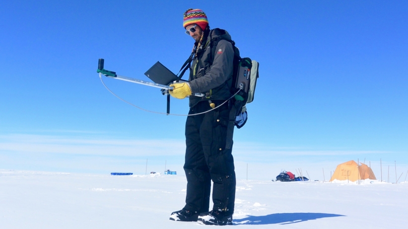 Photograph of researcher Gabe Lewis standing on the snowy surface of the Greenland Ice Sheet holding a computer and scientific instruments, with tents and a snowmobile in the background.