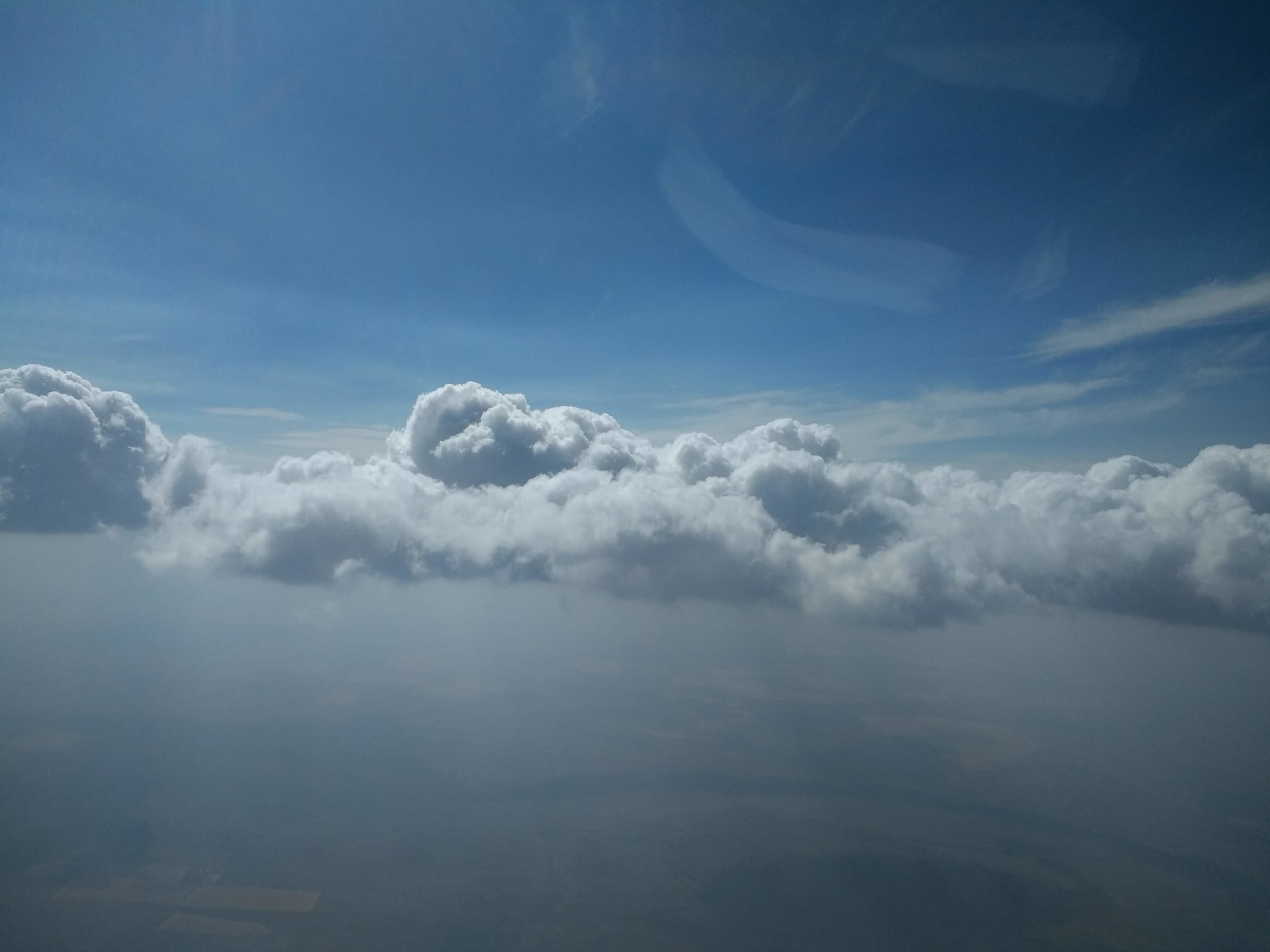 A layer of gray smoke hangs over green and brown mountains, with trees and fields below. White clouds intermingle with the smoke, and the sky is blue but hazy above the clouds. A bit of plane is visible in the image’s center left.