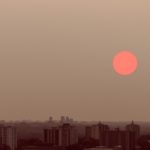 The sun is a matte, neon pink in a hazy brown sky over a smoky city skyline.