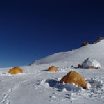 A cluster of orange tents in deep snow on a mountainside, with a blue sky.