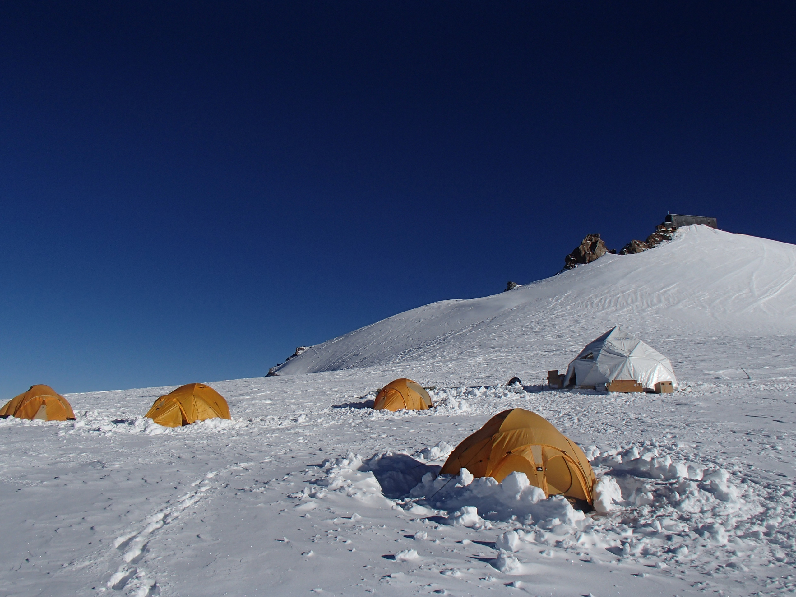 A cluster of orange tents in deep snow on a mountainside, with a blue sky.