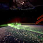 Photograph features green ribbons of the Southern Lights seen from low Earth orbit over the Southern Ocean, framed by solar array and modules of the International Space Station.