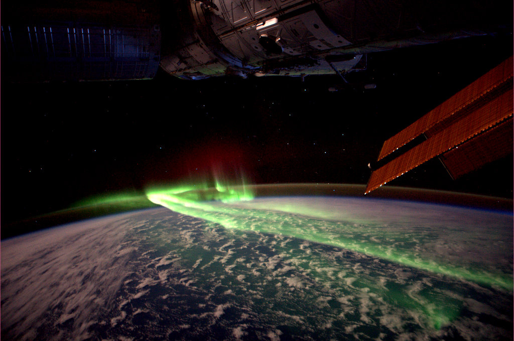 Photograph features green ribbons of the Southern Lights seen from low Earth orbit over the Southern Ocean, framed by solar array and modules of the International Space Station.