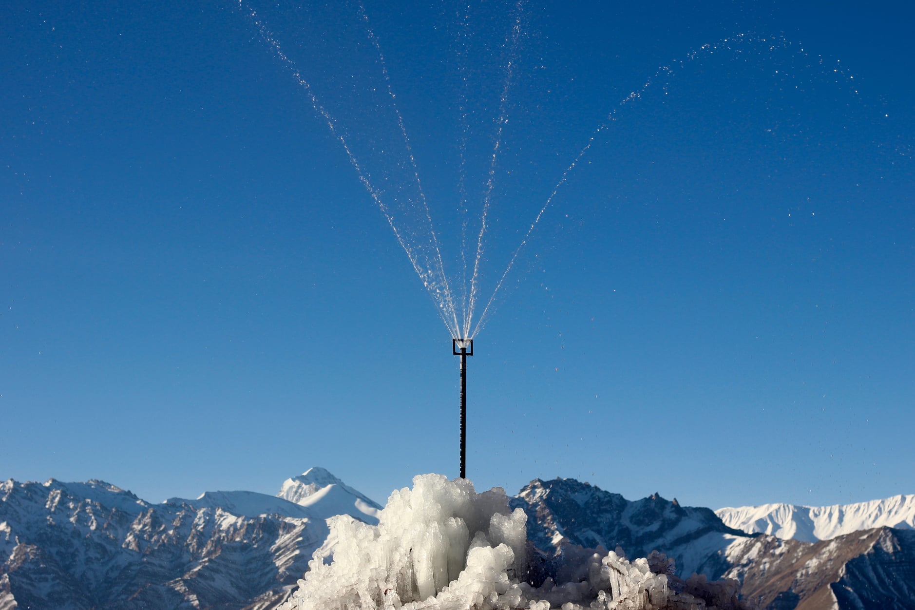 A sprinkler head on a tall pipe sprays five thin streams of water upward against a blue sky. Ice clusters around the base of the fountain. Snowy mountain peaks are in the background. 