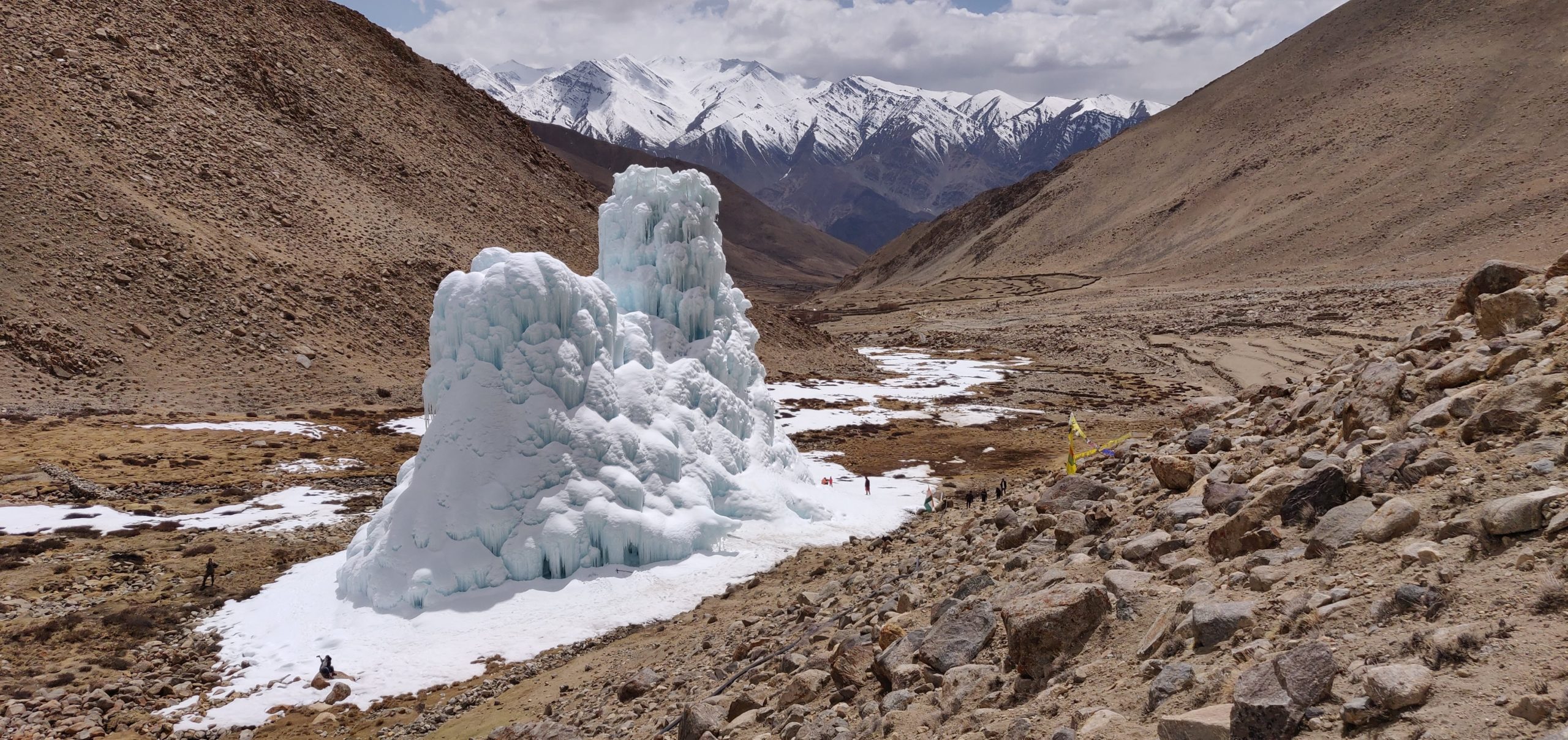 Two ice towers sit in a dry, brown valley of stone, soil, and sparse winter vegetation, with snowy mountain peaks in the background. People appear as tiny figures standing around the edges of the towers.