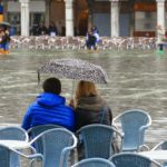 Two people sit on chairs under an umbrella, looking out at a flooded square in a city.