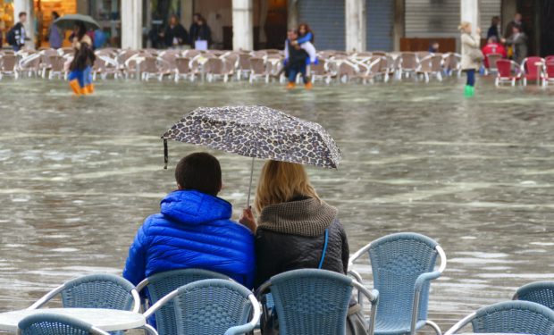 Two people sit on chairs under an umbrella, looking out at a flooded square in a city.