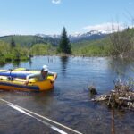 A researcher helps launch a yellow inflatable raft holding a second researcher on a shallow pond surrounded by trees and low shrub, with mountains in the background. Two strings and a measuring tape are strung across the pond about a yard above the water surface.