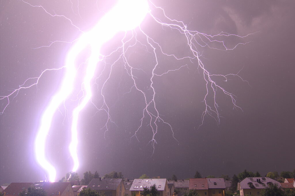 A large and bright lightning bolt flashes against a night sky over a neighborhood.