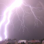 A large and bright lightning bolt flashes against a night sky over a neighborhood.