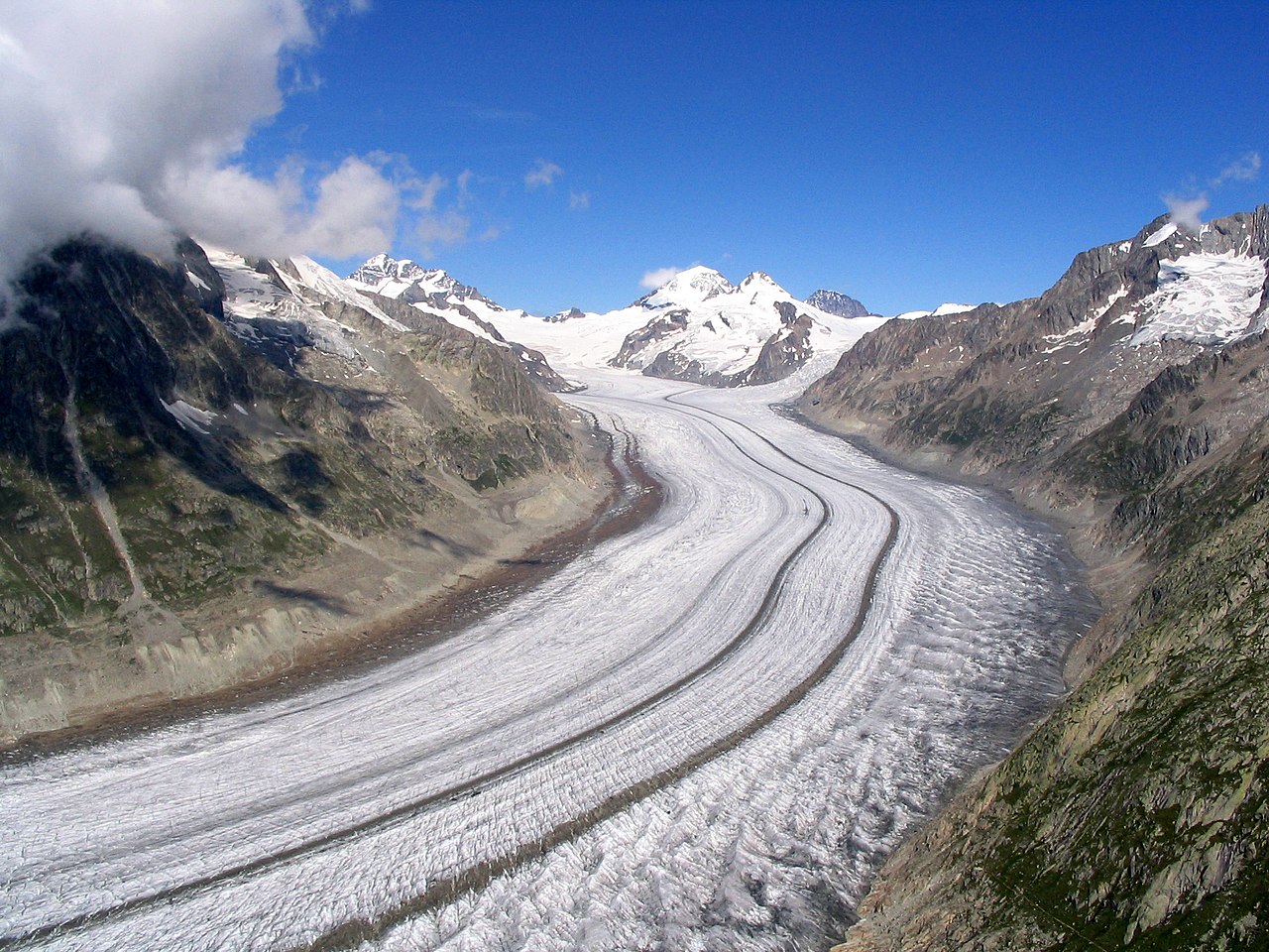Frozen river of a the Aletsch Glacier curves in the forground. The peaks of Jungfrau, Monch and Trogberg are in the background against a blue sky.