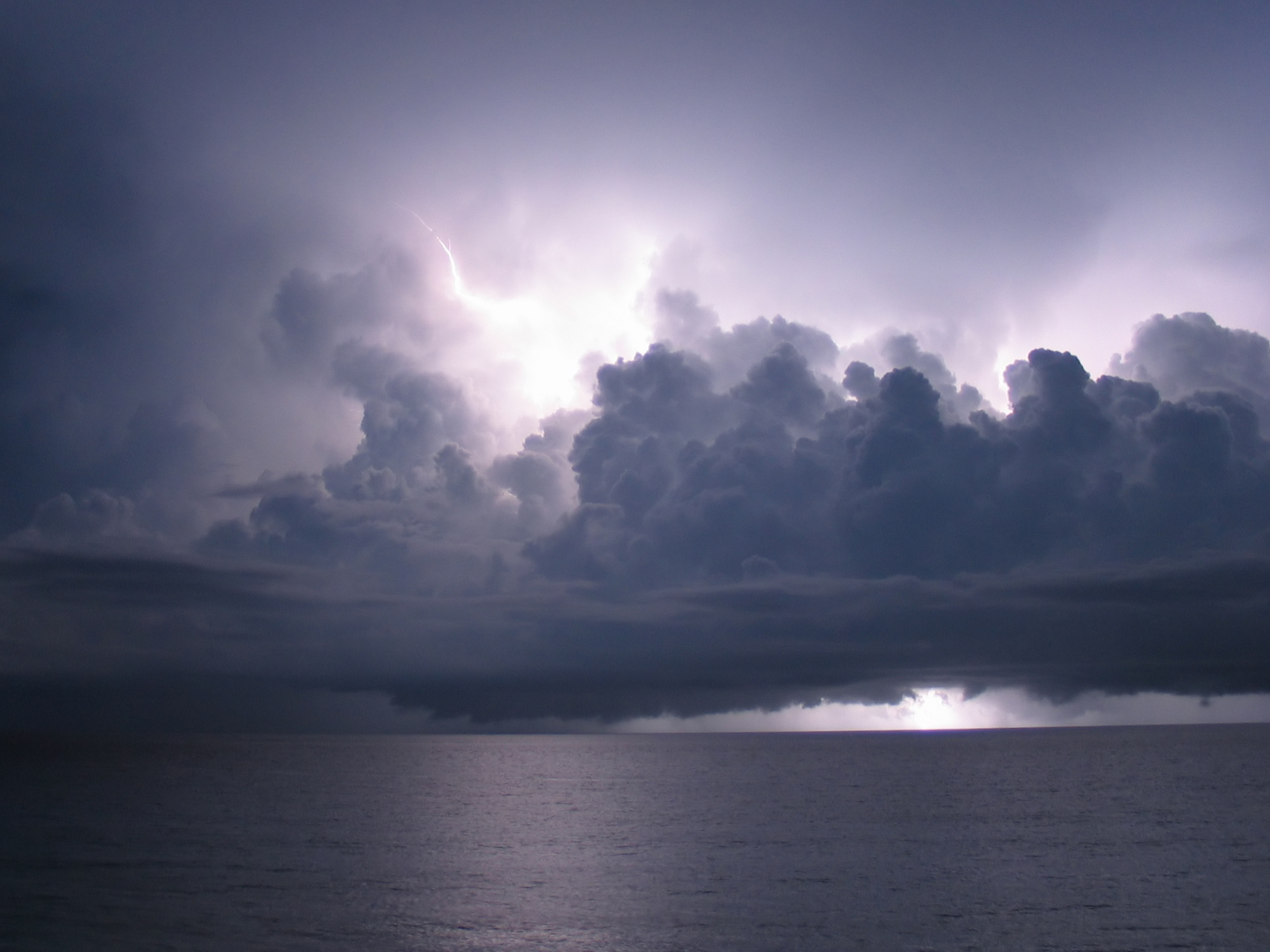 Dark thunderclouds are lit from within by lightning over a flat grey ocean.