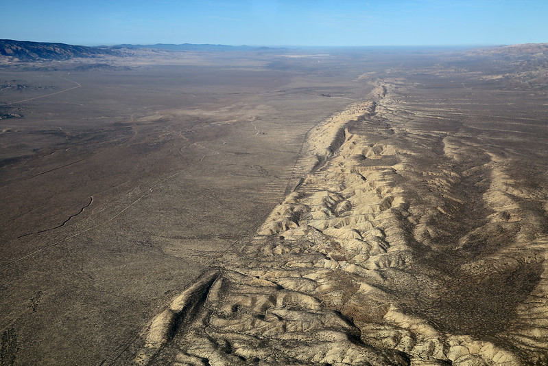 Aerial photograph of a dry grass plain, blue sky in background. A furrow of the San Andreas Fault runs vertically.