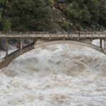 A photograph of a bridge suspended between two rocky hills. Underneath, foamy, brown water rages downstream. A family sits on the bridge watching the water.