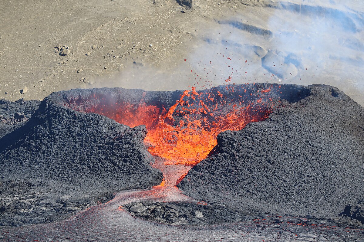 A photograph of a volcano shaped like an ant hill with rising walls, a basin filled with bubbling orange and red lava, and a valley formed in the middle of the photo where the lava is spilling from the volcano. Smoke rises from the lava and partially blocks a tan dirt hill in the background.