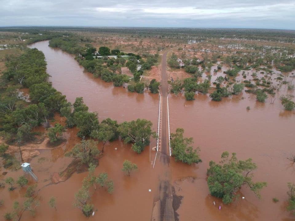 Muddy water flooding over the banks of a river and inundating a road in a wide, flat landscape dotted with tree and shrubs, seen from the air.