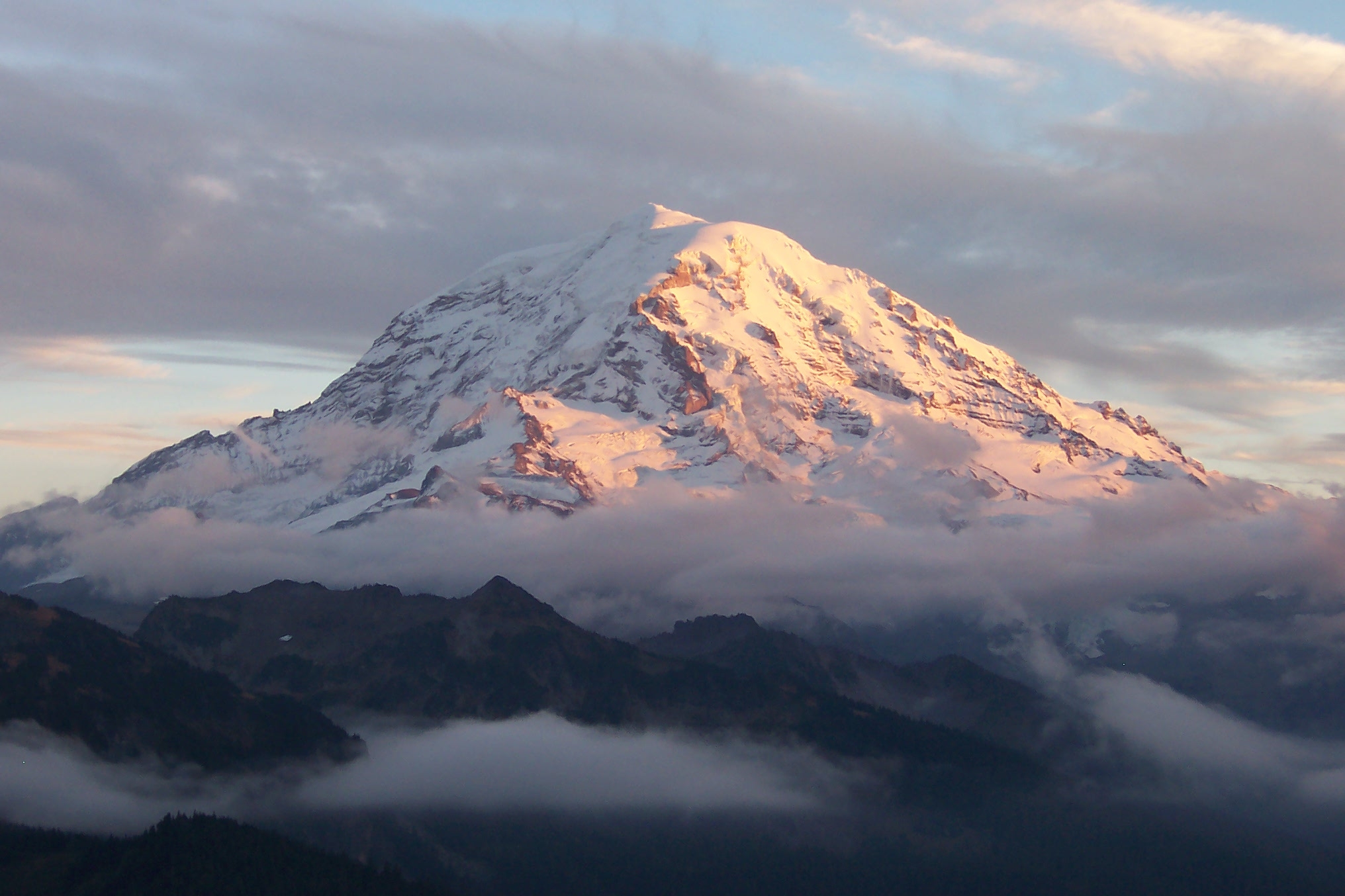A massive, snowy mountain at sunset, wreathed in clouds and towering over smaller, tree-covered mountains below