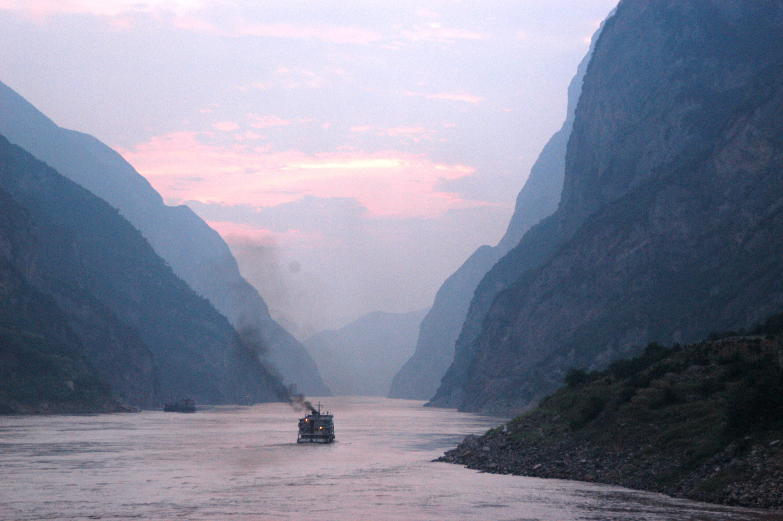 A boat emitting a trail of smoke on a wide, peaceful river between steep cliffs at dusk.