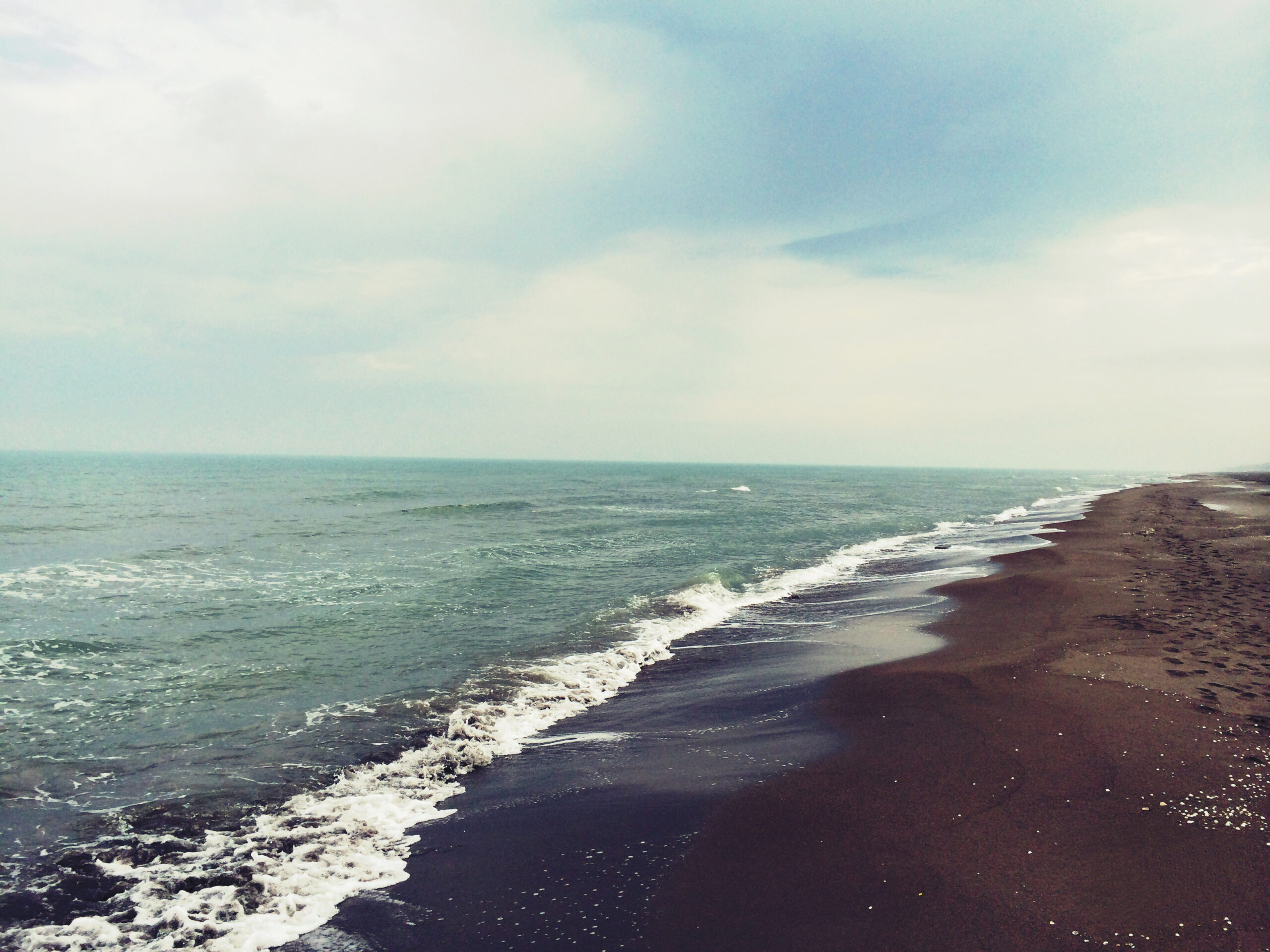 A sandy coastline of a tranquil sea curves off into the distance under a sunny sky with wispy clouds.