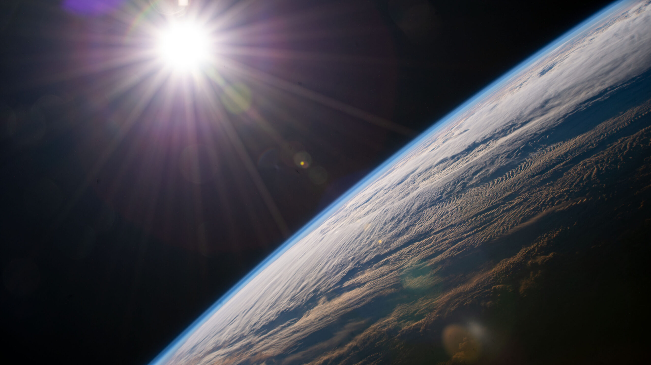 The sun shining over the face of the Earth as seen from space, with Earth's surface in the foreground and the sun in the background