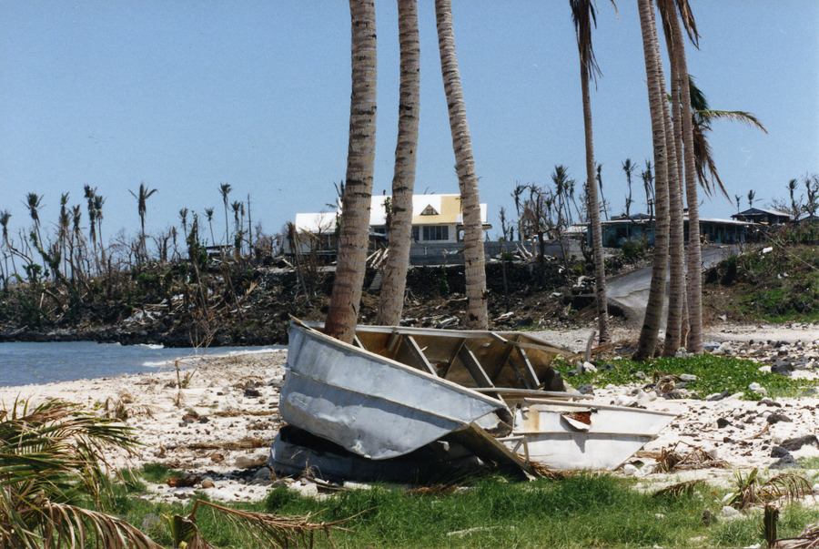 A damaged boat on a beach after a cyclone on a sunny day with trees, coastline and a building in the background.