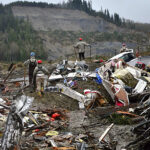 Four people in hard hats walking on a pile of mud, fragments of buildings, and other debris in front of a hill covered in mud with all vegetation stripped away in a rectangular area.