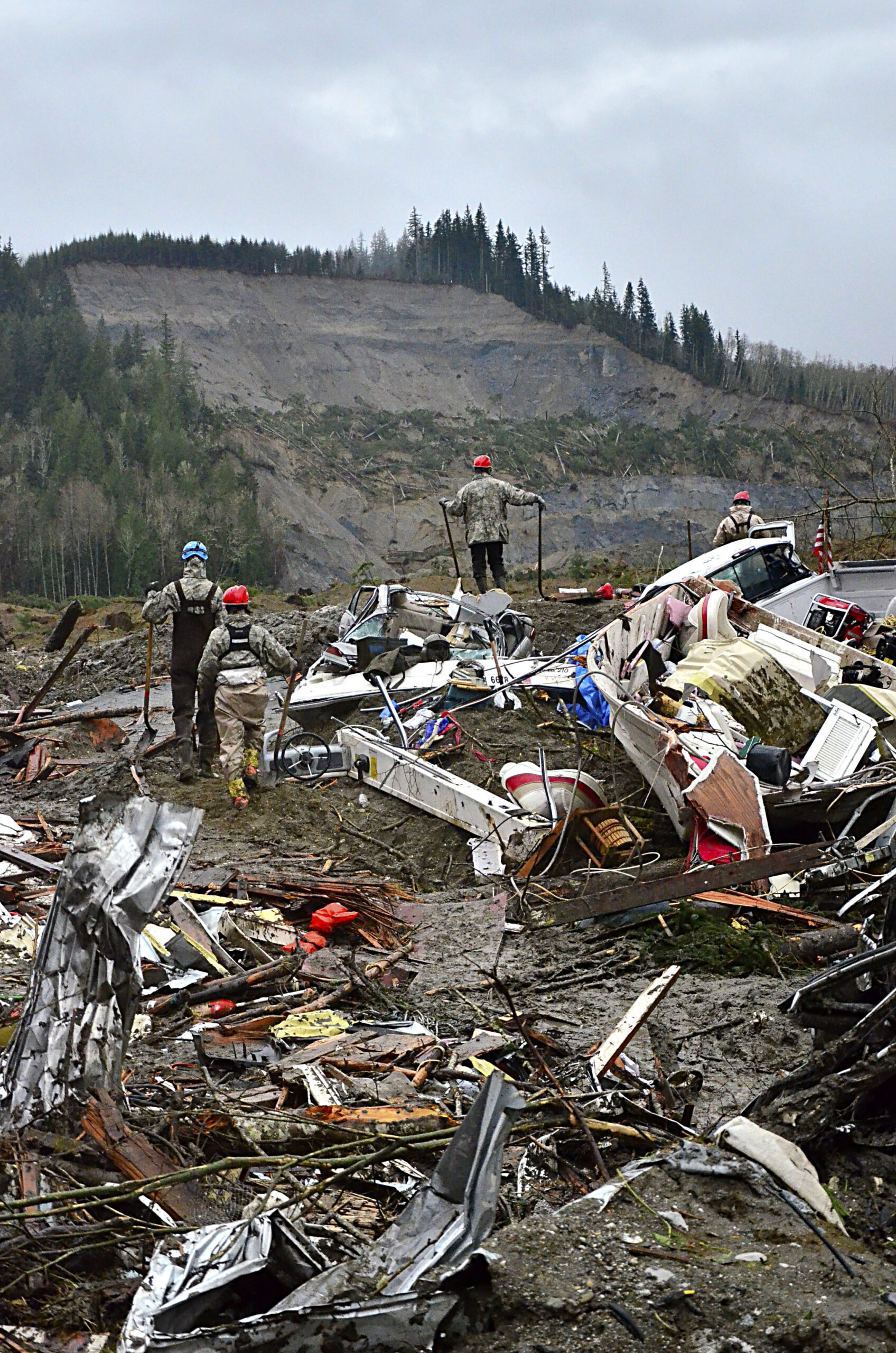 Four people in hard hats walking on a pile of mud, fragments of buildings, and other debris in front of a hill covered in mud with all vegetation stripped away in a rectangular area.