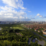 An aerial view of a sprawling city park separated from a residential area by a busy road, with high-rises and skyscrapers in the background.