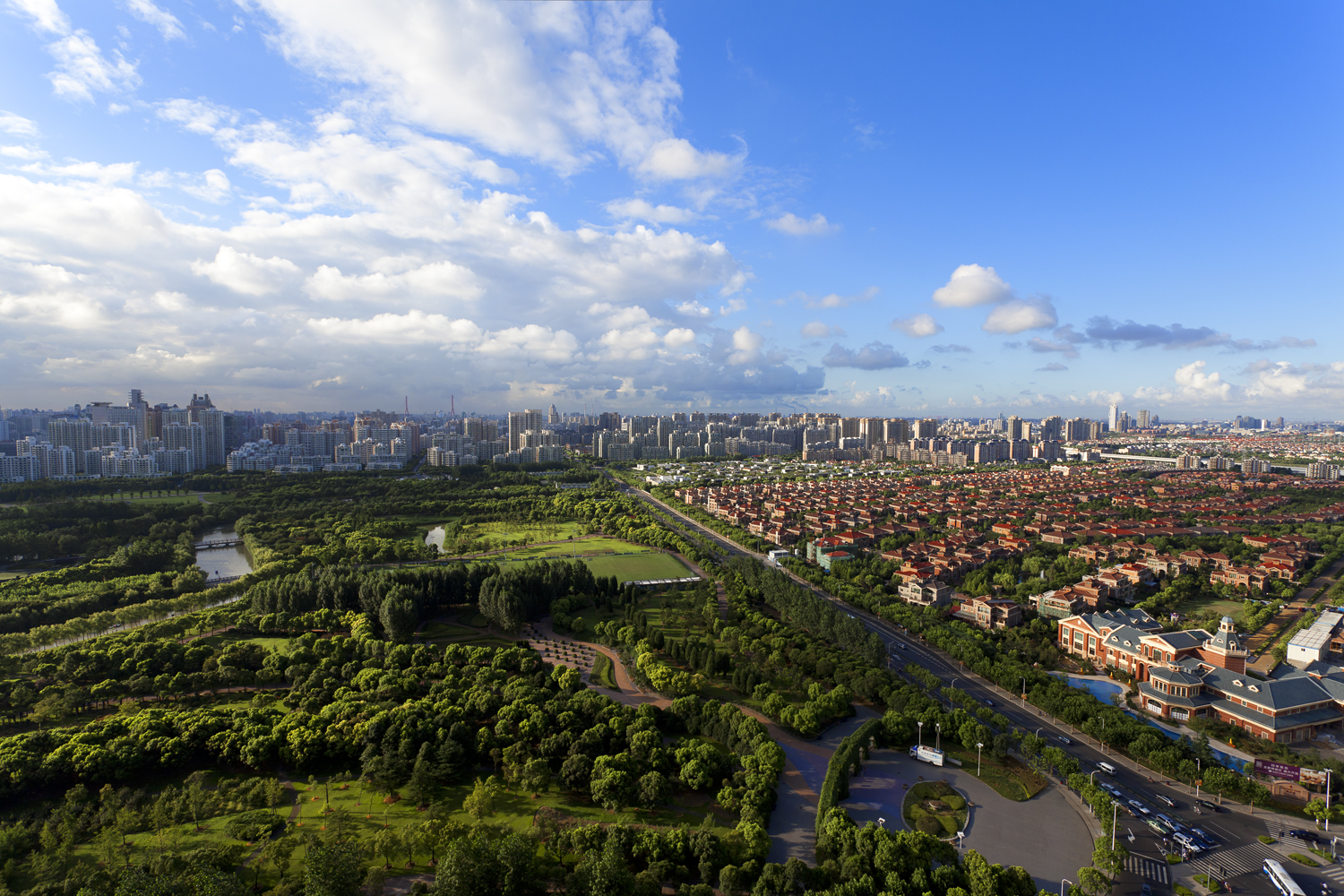 An aerial view of a sprawling city park separated from a residential area by a busy road, with high-rises and skyscrapers in the background.