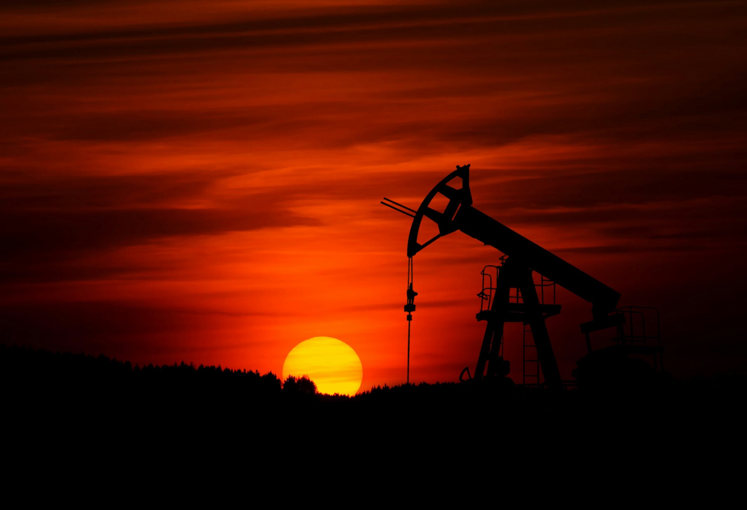 The dark silhouette of an oil derrick in the foreground and a setting sun in the background
