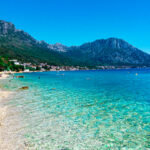 A sunny Croatian beach scene along crystal-clear Adriatic waters, with beachgoers, a beachfront town, and mountains visible in the background.