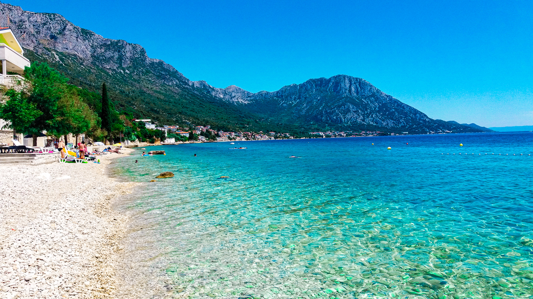 A sunny Croatian beach scene along crystal-clear Adriatic waters, with beachgoers, a beachfront town, and mountains visible in the background.