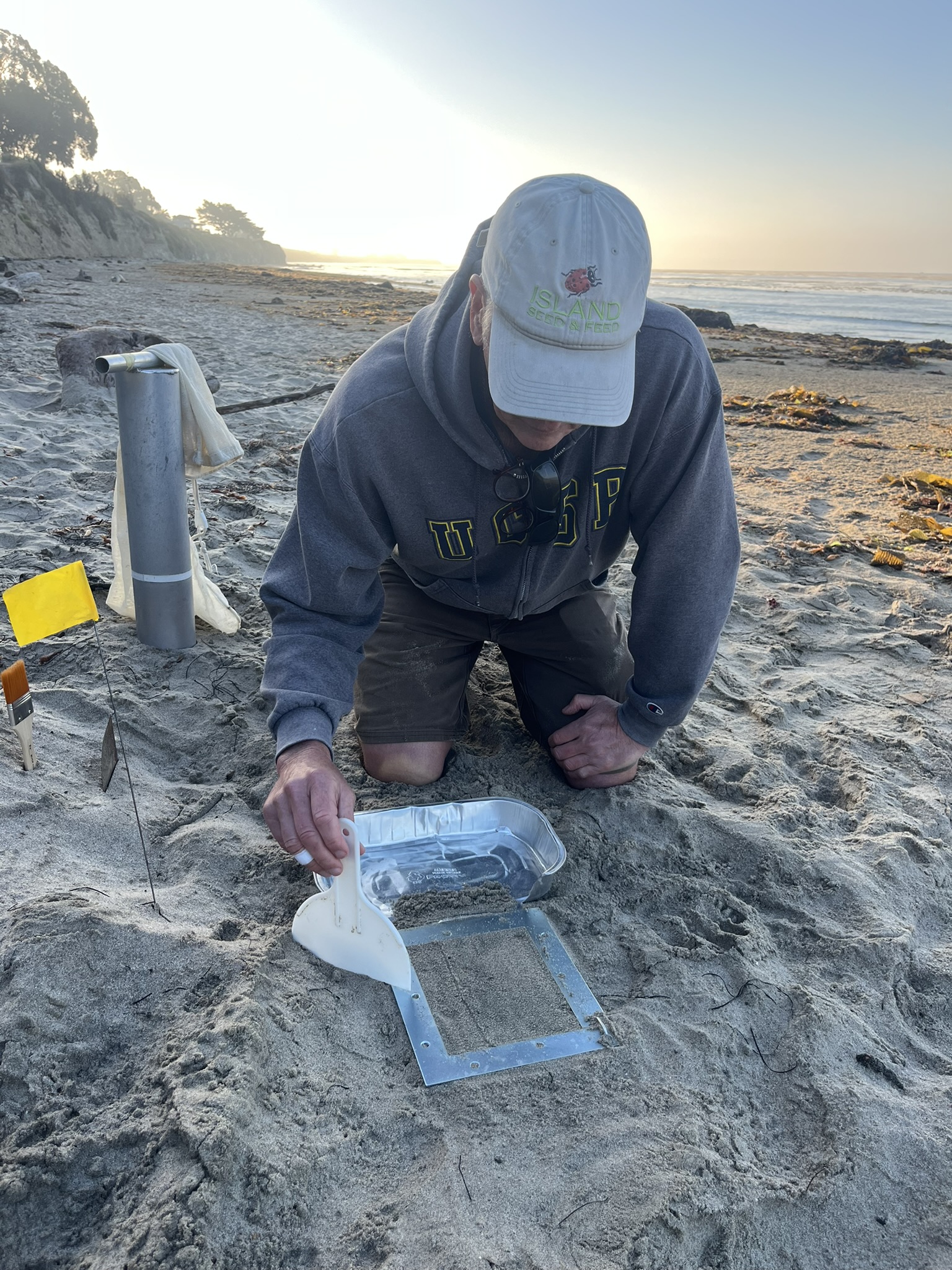 A man in a baseball cap on the beach uses a scraping tool to collect mounded sand in a container.