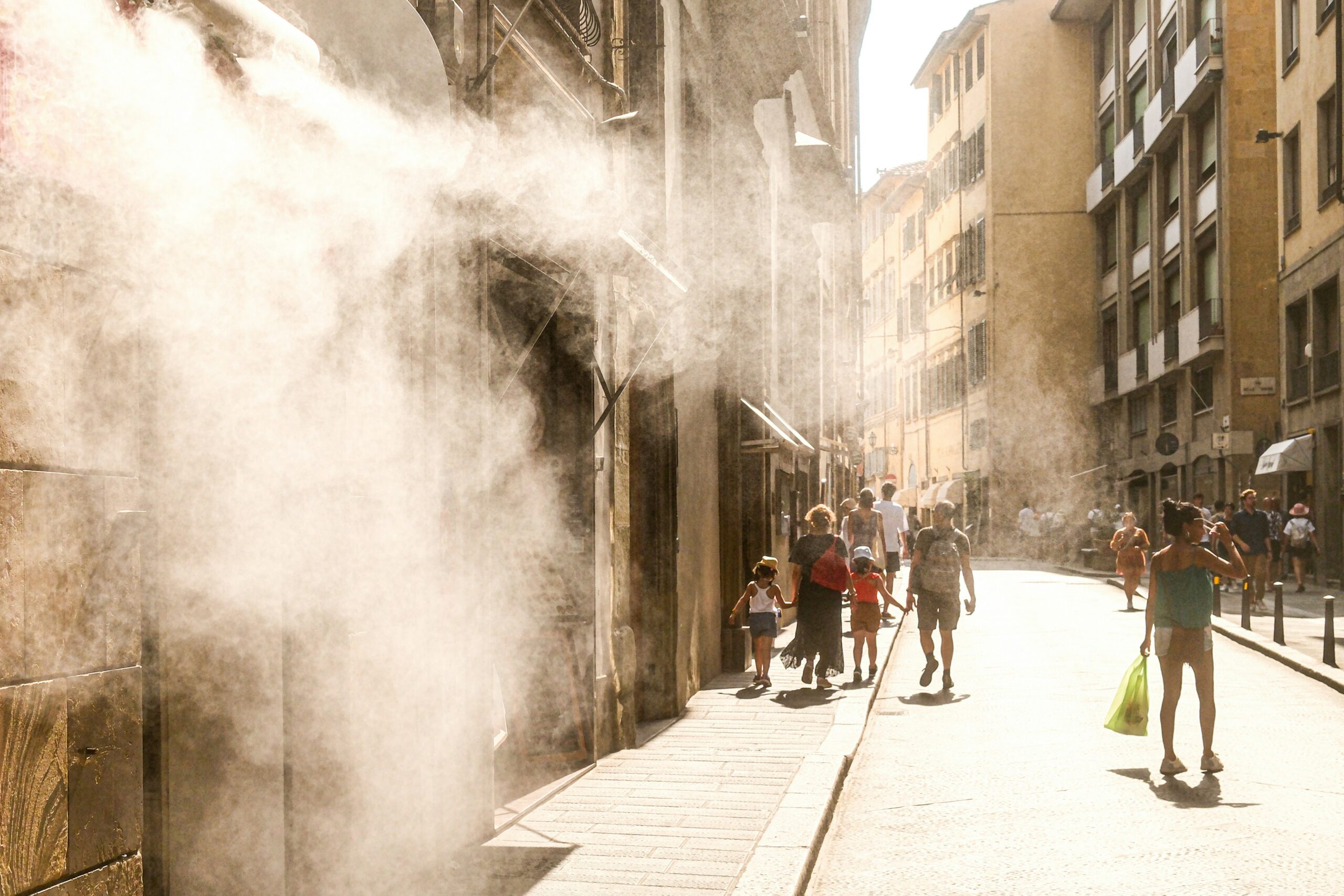 A hot, sunny street in Italy with pedestrians walking under cooling water mist.