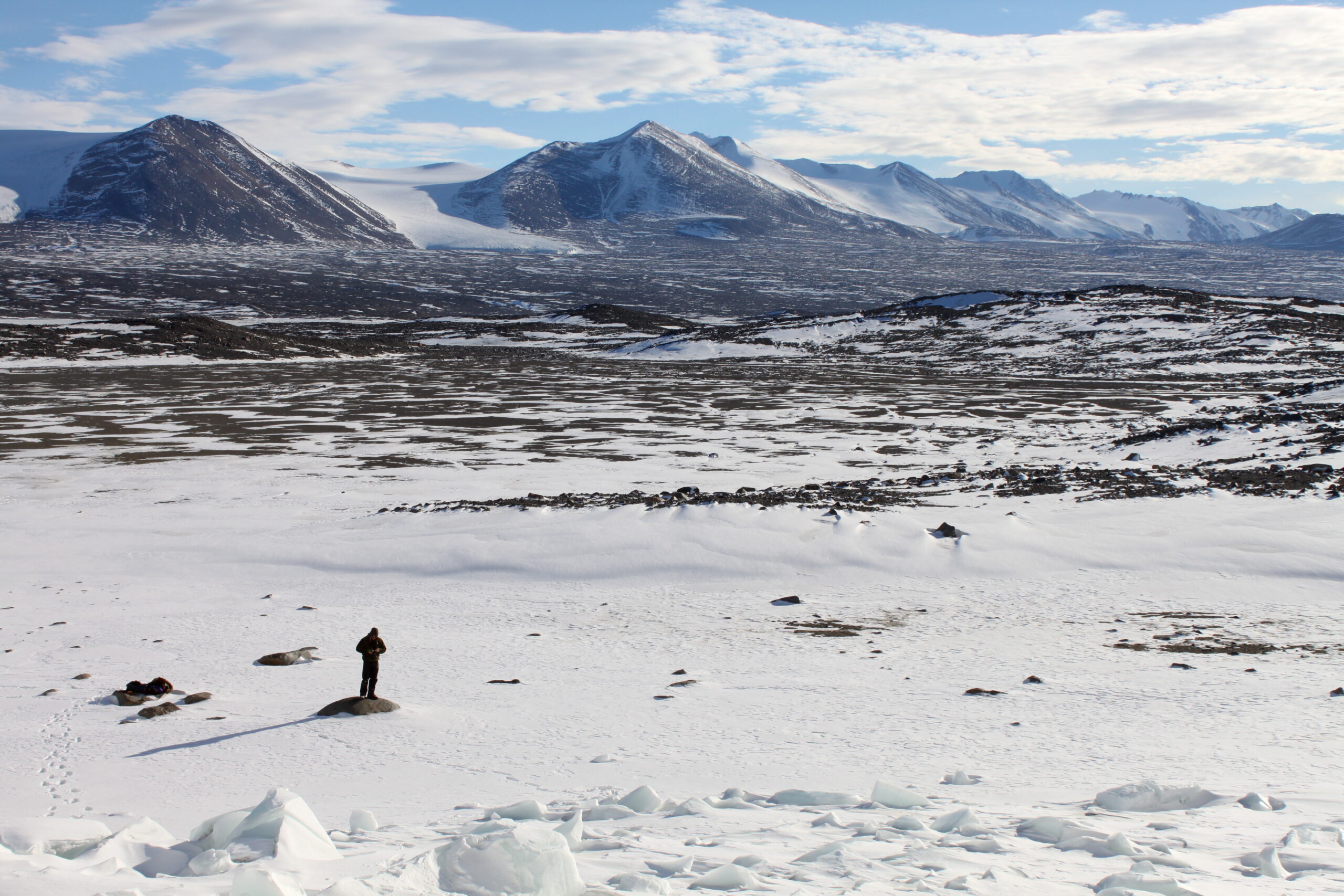 A lone figure standing in the bed of a snowy Antarctic valley on a sunny day, with low mountains in the backdrop