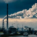Cars driving past steaming smokestacks against a cloudy gray sky.
