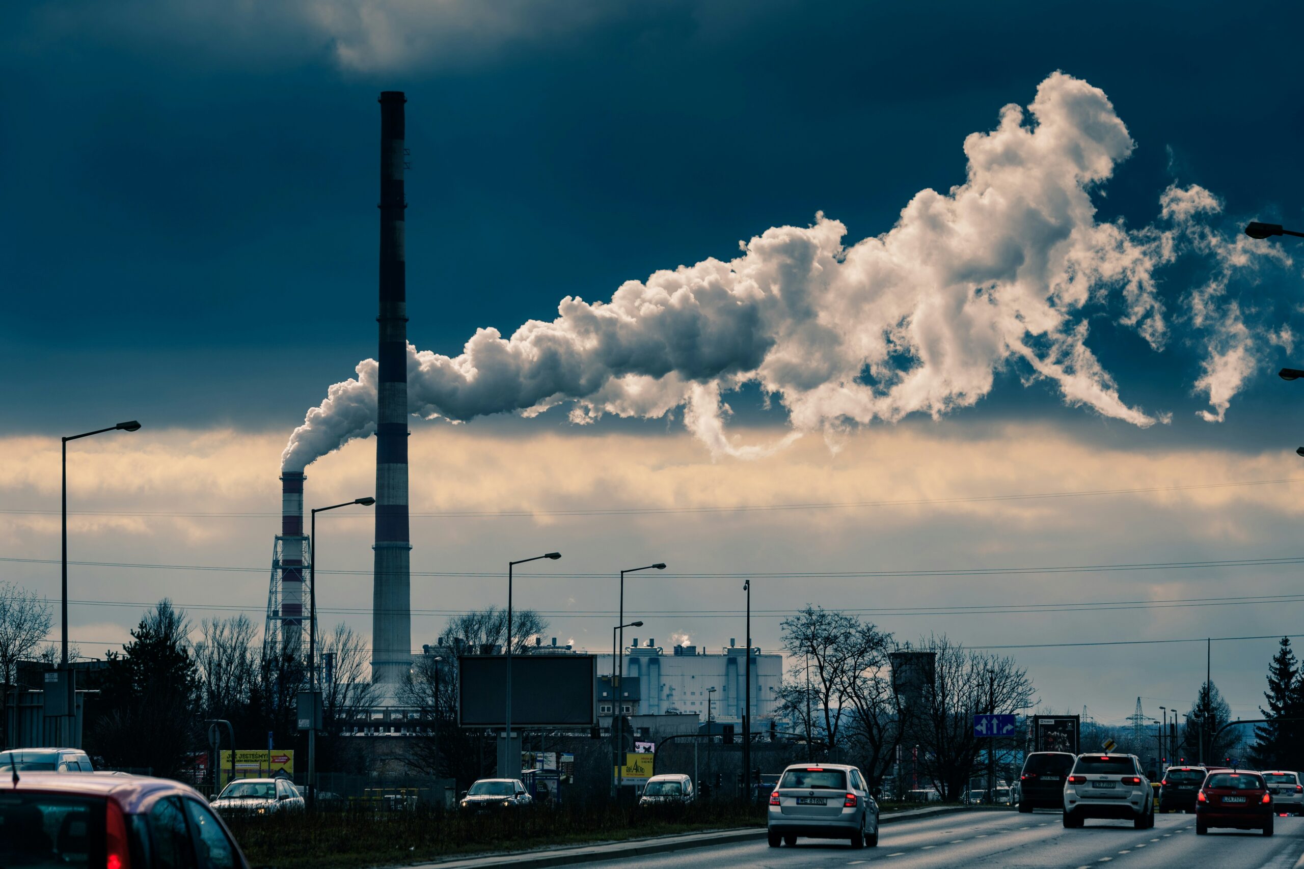Cars driving past steaming smokestacks against a cloudy gray sky.